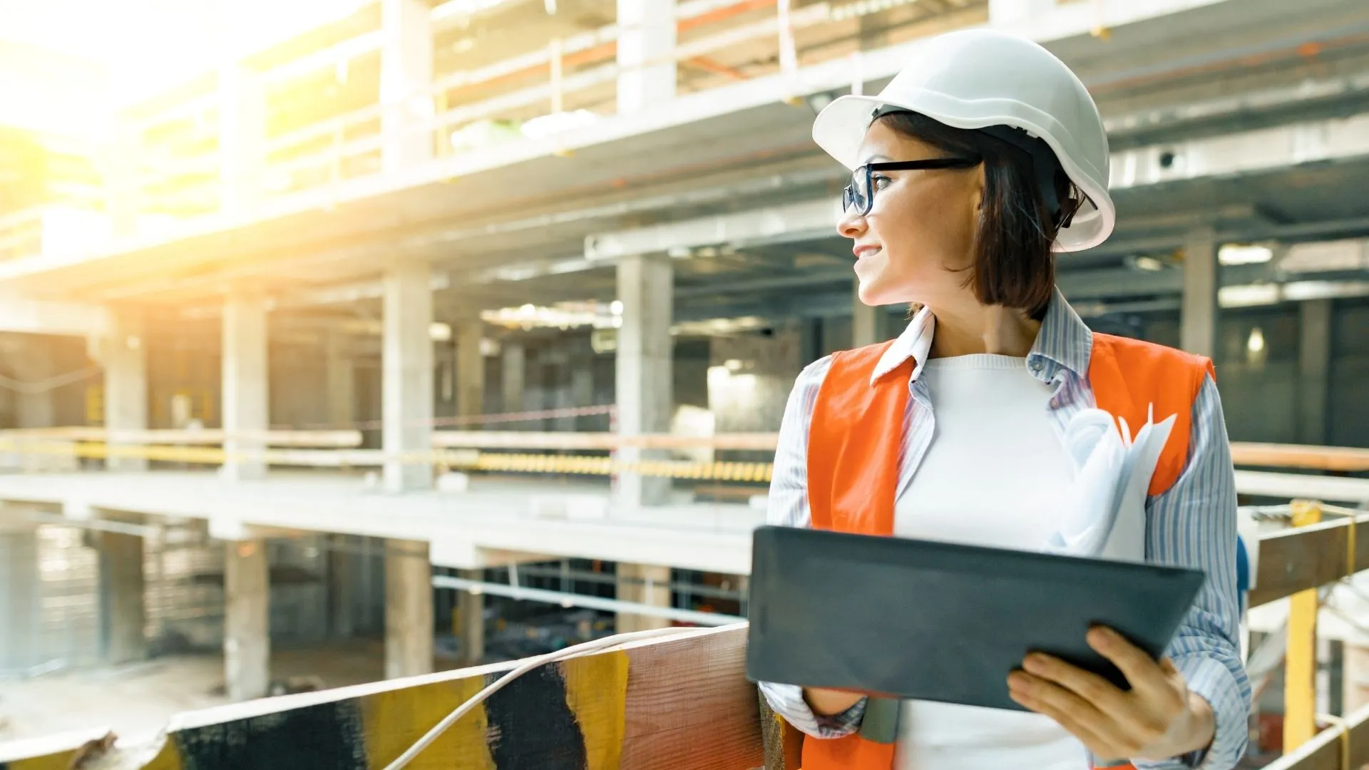 Civil engineer in an orange vest holding a computer, looking away from the camera at an underconstruction building site.